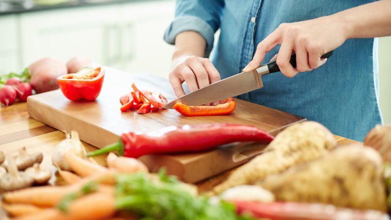 Frau in Jeanshemd steht in der Küche und schneidet eine rote Paprika auf einem Holzbrett auf