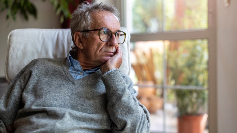 Ein älterer Mann mit Brille sitzt nachdenklich in einem Sessel und blickt aus dem Fenster in einem hellen Raum mit Pflanzen im Hintergrund.
